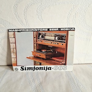 Op de afbeelding: Een vintage advertentie voor een houten stereoconsole. De console heeft een radio, platenspeler en opbergruimte. De advertentie heeft de tekst "MASHPRIBORINTORG SSSSR - MOSKVA" en "Simfonija-003".