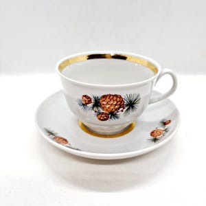 May include: A white porcelain teacup and saucer set with a gold rim and a pine cone design. The cup and saucer have a delicate floral pattern with brown pine cones and green pine needles.