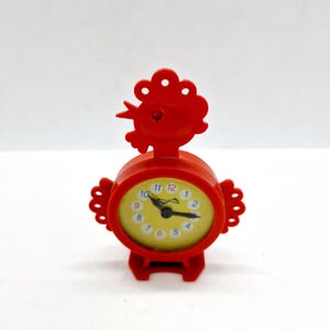 Könnte beinhalten: Eine kleine, rote Uhr in Form eines Hahns mit gelbem Zifferblatt. Die Uhr hat ein rundes Zifferblatt mit schwarzen Zeigern und weißen Zahlen. Die Uhr ist aus Kunststoff und hat einen kleinen Sockel.