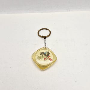 Può includere: Portachiavi vintage con un personaggio di lupo dei cartoni animati. Il lupo ha una testa marrone, capelli blu e una sciarpa rossa, su uno sfondo color crema all'interno di un involucro di plastica trasparente a forma di diamante. Il portachiavi ha un anello di metallo.