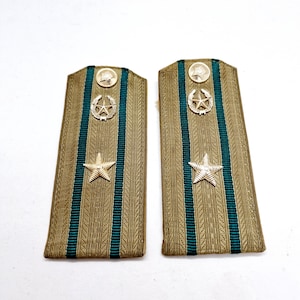 Op de afbeelding: Een paar vintage militaire schouderstukken. De schouderstukken zijn voornamelijk bruin met een blauwgroene streep in het midden. Elk schouderstuk heeft een gouden ster, een lauwerkrans embleem en een cirkelvormig embleem aan de bovenkant.