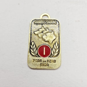 以下が含まれることがあります： 上部に「SKIRNU SKATE」の文字が入った、金色の金属製ペンダント。ペンダントには2匹の犬の横顔、白い「I」が入った赤い円、そして「PSAB un DZAB RIGA」の文字が描かれています。