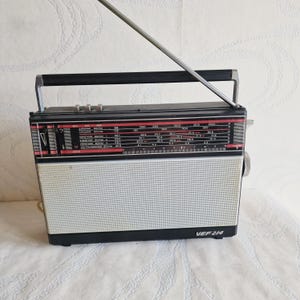 Könnte beinhalten: Ein schwarz-weißes VEF 214 Kofferradio mit rechteckigem Gehäuse, einem Tragegriff an der Oberseite und einer ausgefahrenen Metallantenne. Die Front zeigt eine Frequenzskala mit kyrillischer Beschriftung und ein großes Lautsprechergitter.