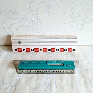 May include: A teal plastic drafting ruler with a clear edge and a sliding center piece, featuring Cyrillic text and a scale. It is accompanied by a rectangular white cardboard box with black horizontal lines and the Cyrillic text REISHINA.