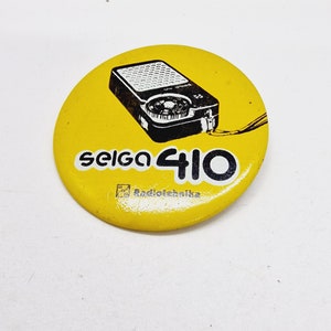 May include: A yellow round pin with a black and white image of a vintage radio with the text "Seiko 410" and "Radiotehnika" printed on it.