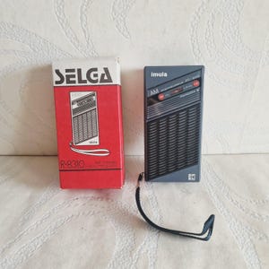 Könnte beinhalten: Ein grauer tragbarer Radioempfänger mit schwarzem Lautsprechergitter und schwarzer Handschlaufe. Das Radio trägt die Aufschrift imula und AM auf der Vorderseite. Es wird neben seiner originalen rot-weißen Pappschachtel mit der Aufschrift SELGA R-8310 gezeigt.