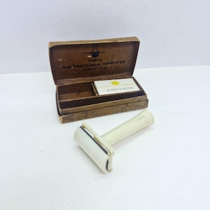 May include: A vintage off-white plastic safety razor with a ribbed handle, accompanied by a rectangular brown cardboard box. The box lid features a black graphic and Cyrillic text, while the interior contains a small white blade case.