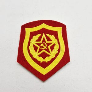 Peut inclure: Écusson militaire rouge et jaune. Il présente une étoile jaune avec un symbole de marteau et de faucille, entourée d'une couronne de laurier et d'une bordure jaune. De forme de bouclier.