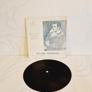 Könnte beinhalten: Eine schwarze Schallplatte mit einem weißen Label, auf dem "Melodiya" steht und ein Porträt eines Mannes abgebildet ist. Die Schallplatte liegt auf einer weißen Oberfläche mit einem weißen Papierumschlag, auf dem "Muslum Magomayev" und "Gibkaya Grammplastinka" steht.