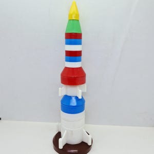 May include: A vertical toy rocket constructed from stackable plastic segments in yellow, green, red, blue, and white. The structure features a pointed yellow nose cone, a cylindrical body with alternating color bands, white fins, and a brown circular base.