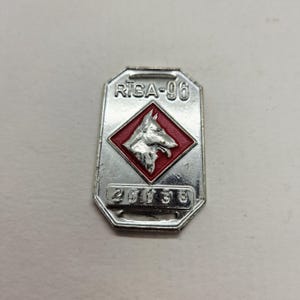 以下が含まれることがあります： 上部に「RIGA-96」、下部に「20138」と書かれた銀色の金属バッジ。中央の赤いひし形には犬の頭が描かれています。バッジは八角形です。