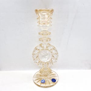 May include: A clear, decorative glass candlestick holder with a round base and a detailed, faceted design. The center features a round emblem with a bird. The candlestick is approximately 10 inches tall.