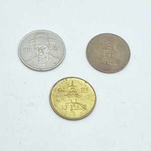 May include: Three South Korean coins: one silver-colored 100 won coin featuring a portrait of a man in a traditional hat, one bronze-colored 10 won coin, and one gold-colored 10 won coin, both featuring a pagoda design and Korean text characters.