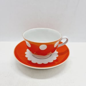 May include: A white teacup with a bright orange saucer. The teacup has a white polka dot pattern on the orange exterior. The saucer has a scalloped edge.