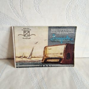 Puede incluir: Manual de instrucciones vintage para una radio, con una escena de veleros y una ilustración de radio retro. La portada está dividida en dos secciones, con texto en ruso y el año 1955 en la parte inferior. El manual está sobre una superficie blanca.