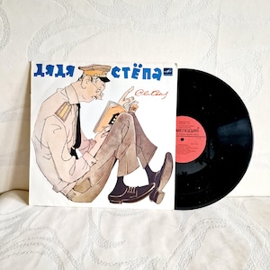 May include: A vintage vinyl record featuring a cover illustration of a man in uniform reading a book. The cover has Russian text and a cartoon-style drawing. The record itself is black with a red label.