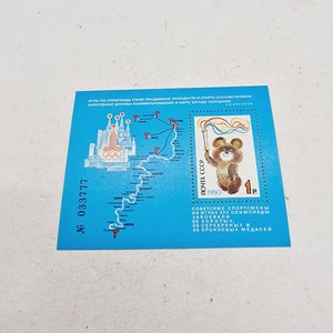 Può includere: Un francobollo blu e bianco con una mappa del percorso della torcia olimpica per le Olimpiadi estive del 1980 a Mosca, URSS. Il francobollo presenta un orso dei cartoni animati che tiene una torcia e il testo "ПОЧТА СССР 1980 1р".