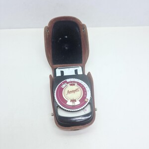 May include: A vintage Leningrad 2 light meter housed in a brown leather case. The device features a circular dial with a maroon and white face, displaying numerical scales and Cyrillic text. The black plastic body is secured within the open case.