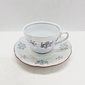 May include: A white porcelain teacup and saucer set with a floral design in gold and blue. The cup has a scalloped rim and the saucer has a scalloped edge.