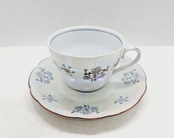 Peut inclure: Une tasse à thé et une soucoupe en porcelaine blanche avec un motif floral en or et bleu. La tasse a un bord festonné et la soucoupe a un bord festonné.