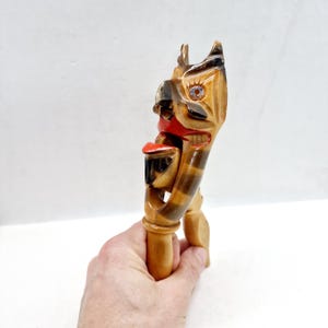 Op de afbeelding: Een gesneden houten notenkraker in de vorm van een gestileerd gezicht. De notenkraker heeft een rode mond, zwart-bruine gestreepte details en een lichtbruine afwerking. Het ontwerp wordt in een hand gehouden.