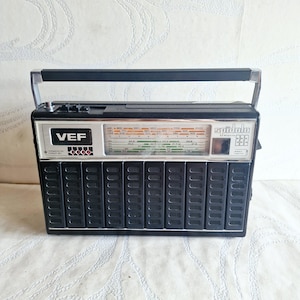 Könnte beinhalten: Ein schwarzes tragbares Radio vom Typ VEF Spidola 232 mit einer silbernen Bedienblende inklusive Frequenzskalen, einem schwarzen Lautsprechergitter mit vertikalen Lamellen und einem Metalltragegriff. Vorne sind das VEF-Logo und Spidola 232.