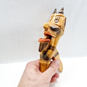 Op de afbeelding: Een gesneden houten notenkraker in de vorm van een grillig gezicht met een open mond. De notenkraker heeft een lichtbruine basiskleur met donkerbruine strepen en oranje accenten. De ogen zijn ingelegd met een blauwe steen.