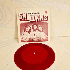 Könnte beinhalten: Eine Vintage-Vinyl-Schallplatte und Albumhülle mit der Band "Bee Gees". Die Albumhülle ist weiß mit rotem Text und einem Foto der Bandmitglieder. Die Schallplatte hat eine einfarbige rote Farbe.