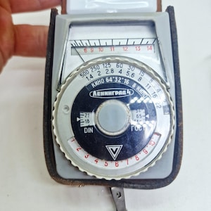 May include: A vintage Leningrad 4 light meter featuring a grey plastic casing and a black circular dial with white numerical markings. The device includes a needle indicator, various aperture settings, and Cyrillic text printed on the face.