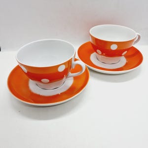 May include: Two orange teacups with white polka dots and matching saucers. The cups and saucers are made of ceramic.