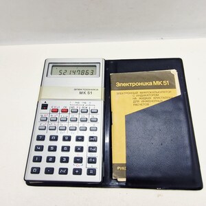 May include: A vintage silver and black Elektronika MK 51 calculator with a black case. The calculator displays the number 52147863. A yellow booklet with Russian text is in the case. The calculator has many buttons for scientific calculations.
