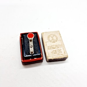 Può includere: Un piccolo interruttore in metallo nero con un pulsante rosso e una leva argentata. L'interruttore è in una scatola rossa e nera con le parole "Series" e "Universal" stampate sulla scatola.