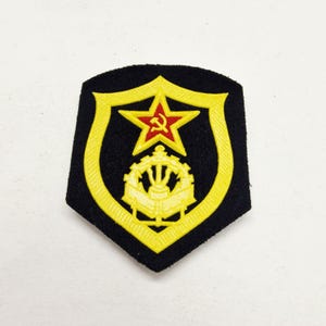 Könnte beinhalten: Ein schwarzer Aufnäher mit gelbem Rand, der einen roten Stern und ein Hammer-und-Sichel-Emblem zeigt. Der Aufnäher hat eine Schildform mit einem gelben Zahnrad-Design am unteren Rand.