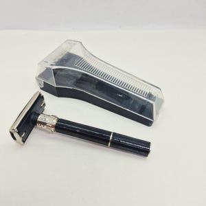 Può includere: Un rasoio di sicurezza nero e argento con una ghiera regolabile che mostra i numeri da 1 a 9, abbinato a una custodia protettiva in plastica trasparente con base nera. Il manico del rasoio ha una texture a coste e una finitura metallica.