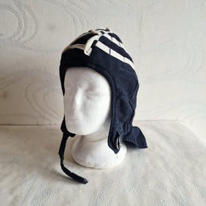 Puede incluir: Gorro de aviador azul marino con rayas blancas y un cordón crema. El gorro tiene orejeras y un detalle de anillo metálico. El gorro se exhibe en una cabeza de maniquí blanca.