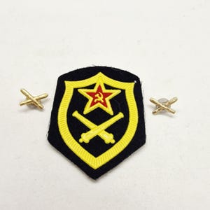 May include: A black and yellow military patch with a red star and hammer and sickle emblem, along with two gold crossed cannons. Two gold crossed cannon pins are also included.