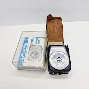 May include: A black and silver light meter in a brown leather case. The light meter is labelled "Leningrad 4" and has a dial with numbers and markings. The case has a strap and a small metal loop.