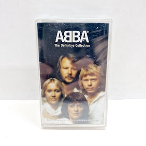 Op de afbeelding: ABBA "The Definitive Collection" cassetteband. De cassettecase heeft een donkere achtergrond met de naam van de band en de albumtitel in het wit. De afbeelding op de cassette toont de vier bandleden.
