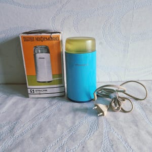 Puede incluir: Un molinillo de café eléctrico de color azul brillante con una tapa amarilla translúcida y un cable de alimentación blanco con enchufe de dos clavijas. El dispositivo viene con su caja de cartón original, que muestra la marca Straume y texto cirílico.