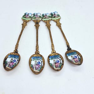 Puede incluir: Un juego de cuatro cucharas de metal dorado con mangos ornamentados y cuencos ovalados. Cada cuchara está decorada con escenas de esmalte de colores que representan a una pareja con vestimenta de época en un jardín, tanto en el mango como en el cuenco.