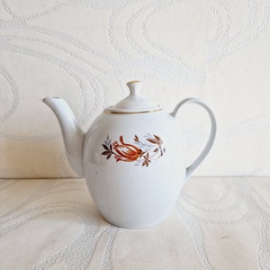 May include: White ceramic teapot with a floral design in shades of orange, brown, and gray. The teapot has a gold trim around the lid and a gold band around the base.
