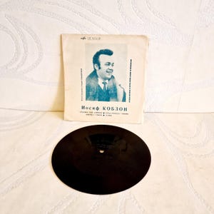 Könnte beinhalten: Eine schwarze Schallplatte mit einem weißen Papierumschlag, der ein Foto eines Mannes mit dem Text "Иосиф КОБЗОН" in Blau zeigt.