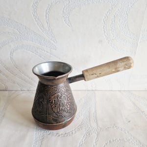 May include: A copper-colored Turkish coffee pot, also known as a cezve, with a wooden handle. The pot features an embossed design of grapes and leaves. The pot is filled with dark liquid, likely coffee, and sits on a white surface.