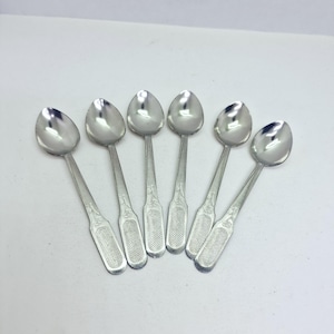 May include: A set of six silver-toned metal spoons arranged in a fan pattern. Each spoon features a smooth, rounded bowl and a flat handle with a textured, cross-hatched rectangular design at the base and a subtle engraved floral motif above.