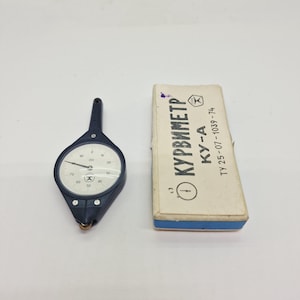 May include: A dark blue map measuring tool with a white circular dial featuring black markings and a needle, accompanied by a rectangular white cardboard box with blue trim, printed with the text KURVIMETR KY-A and TY 25-07-1039-74.