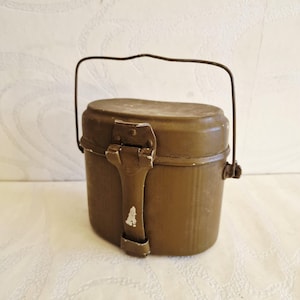 May include: An olive green metal mess kit with a hinged lid and a wire handle. The kit has a rectangular shape with rounded edges and a clasp closure. The paint shows signs of wear, giving it a vintage appearance.