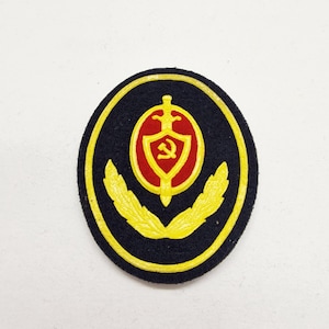 Puede incluir: Parche azul marino con borde amarillo. Presenta un escudo rojo con un emblema de martillo y hoz, rodeado por una espada amarilla y coronado con una corona amarilla. Dos ramas de laurel amarillas en la parte inferior.