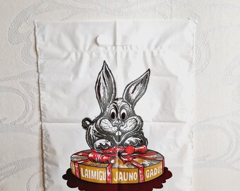 May include: A white plastic shopping bag with a cartoon rabbit holding a red ribbon around a gift box. The gift box is decorated with a gold ribbon and the words "Laimigu Jauno Gadu!" in red and black lettering.
