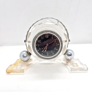 Può includere: Un orologio vintage trasparente simile al cristallo con quadrante nero e dettagli argentati. L'orologio ha una forma sferica con un design sfaccettato e poggia su una base trasparente con piedini a sfera argentati. Il quadrante ha numeri bianchi e lancette rosse.