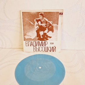 Könnte beinhalten: Eine blaue Schallplatte mit einem weißen Label. Die Schallplatte liegt auf einer weißen Oberfläche. Das Cover der Schallplatte ist im Hintergrund sichtbar. Das Cover ist braun und zeigt ein Foto eines Mannes, der Gitarre spielt. Der Text auf dem Cover lautet "Владимир Высоцкий песни".